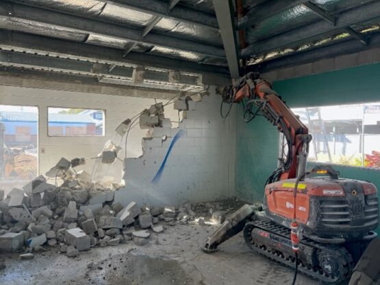 Mini excavator with hydraulic breaker demolishing an internal concrete block wall in a commercial building.